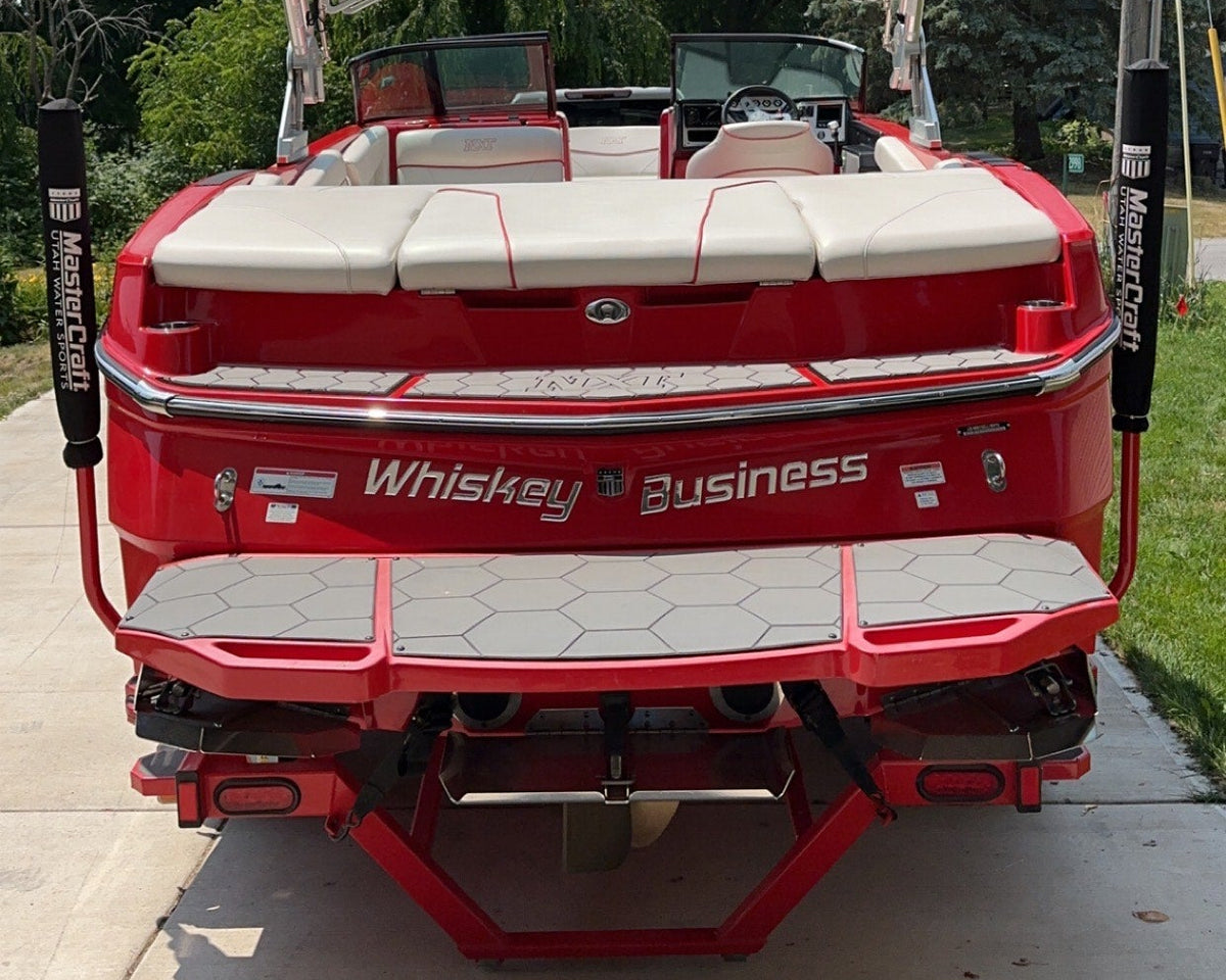 A red and white Mastercraft pontoon boat viewed from the rear, parked outdoors on a concrete surface with trees and utility poles in the background; custom boat name reads Whiskey Business